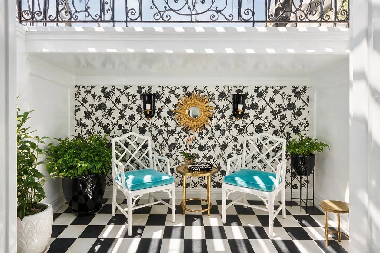 A small outdoor seating nook in Hollywood Regency style with a black-and-white checkered floor, white walls, floral black-and-white wallpaper, and two white lattice-back chairs with bright turquoise cushions. A gold sunburst mirror hangs between black wall sconces, with potted green plants and small brass side tables adding contrast and elegance.
