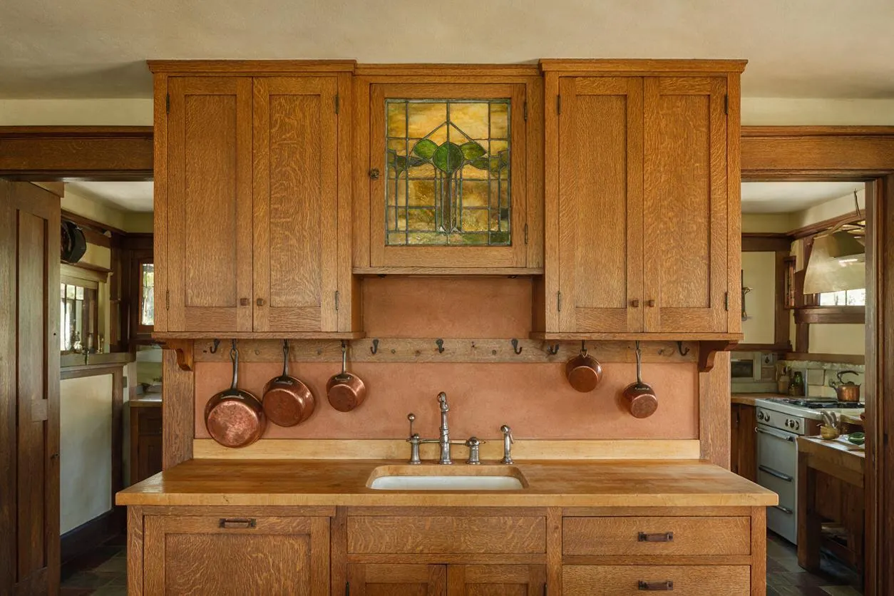 A Craftsman-style kitchen featuring warm oak cabinets, a central sink area, and a stained glass cabinet panel above the backsplash. Copper pots hang from black hooks on a rail, complementing the earthy plaster wall and wood-trimmed doorways. The room has a classic, handcrafted feel with natural finishes and soft, ambient light.