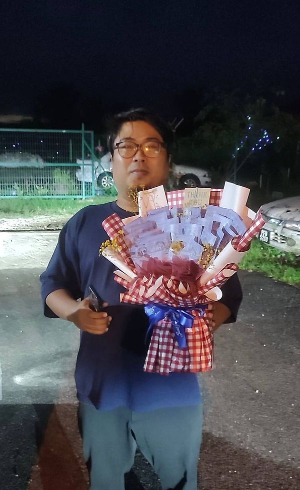 A man holds a decorative bouquet with notes in a dimly lit outdoor setting.