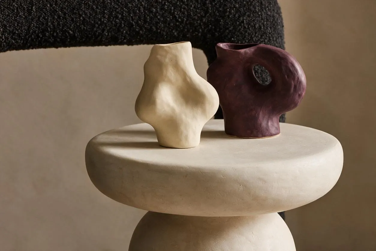A modern decorative vignette shows two small sculptural ceramic vessels on a rounded pedestal table. One vessel is ivory and matte, while the other is deep plum with a cutout handle, set against a warm neutral background and a dark textured textile.