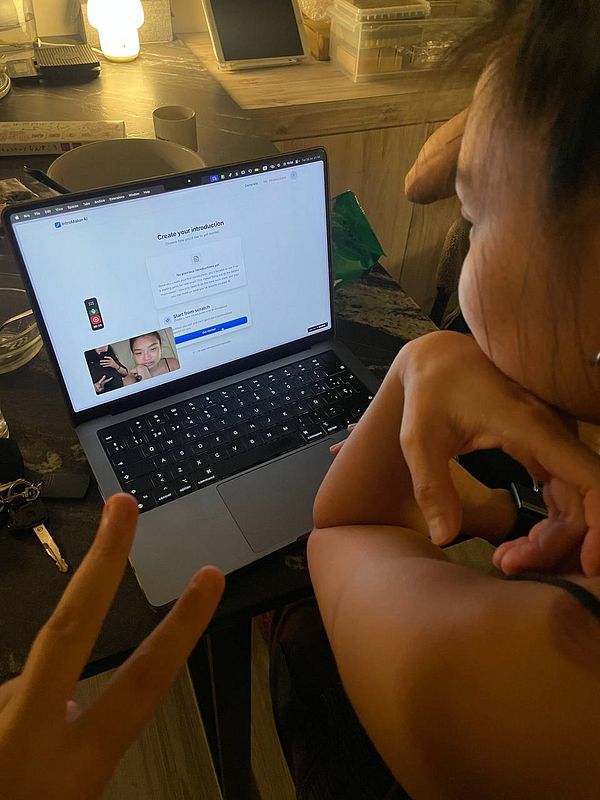 A person is engaged in creating an introduction using IntroMakerAI on a laptop while another individual gives a peace sign in the foreground.