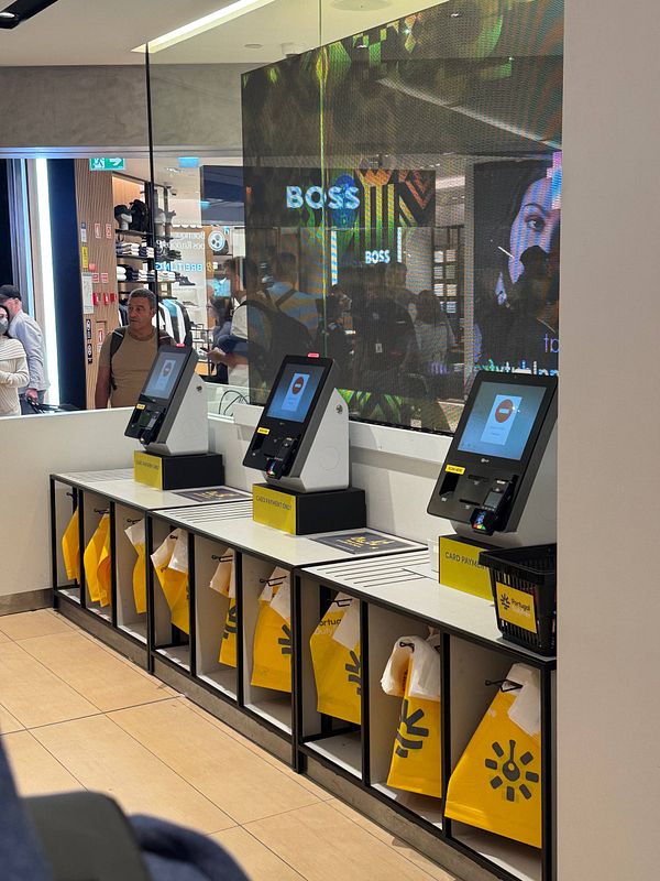 The image depicts a modern self-service kiosk area at Lisbon Airport, featuring multiple payment terminals and storage bins.