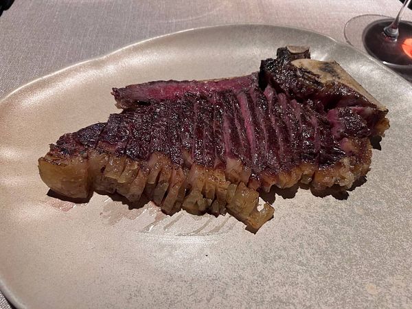 A large grilled monster steak served on a rectangular black plate with some greens on the side and a dark beverage in a glass.