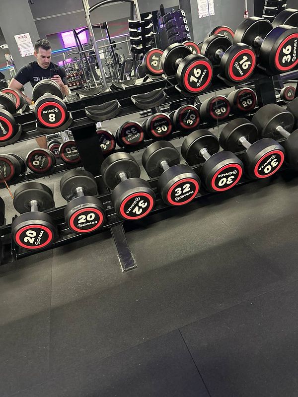 A gym setting featuring a rack of dumbbells and a person taking a selfie in the mirror.