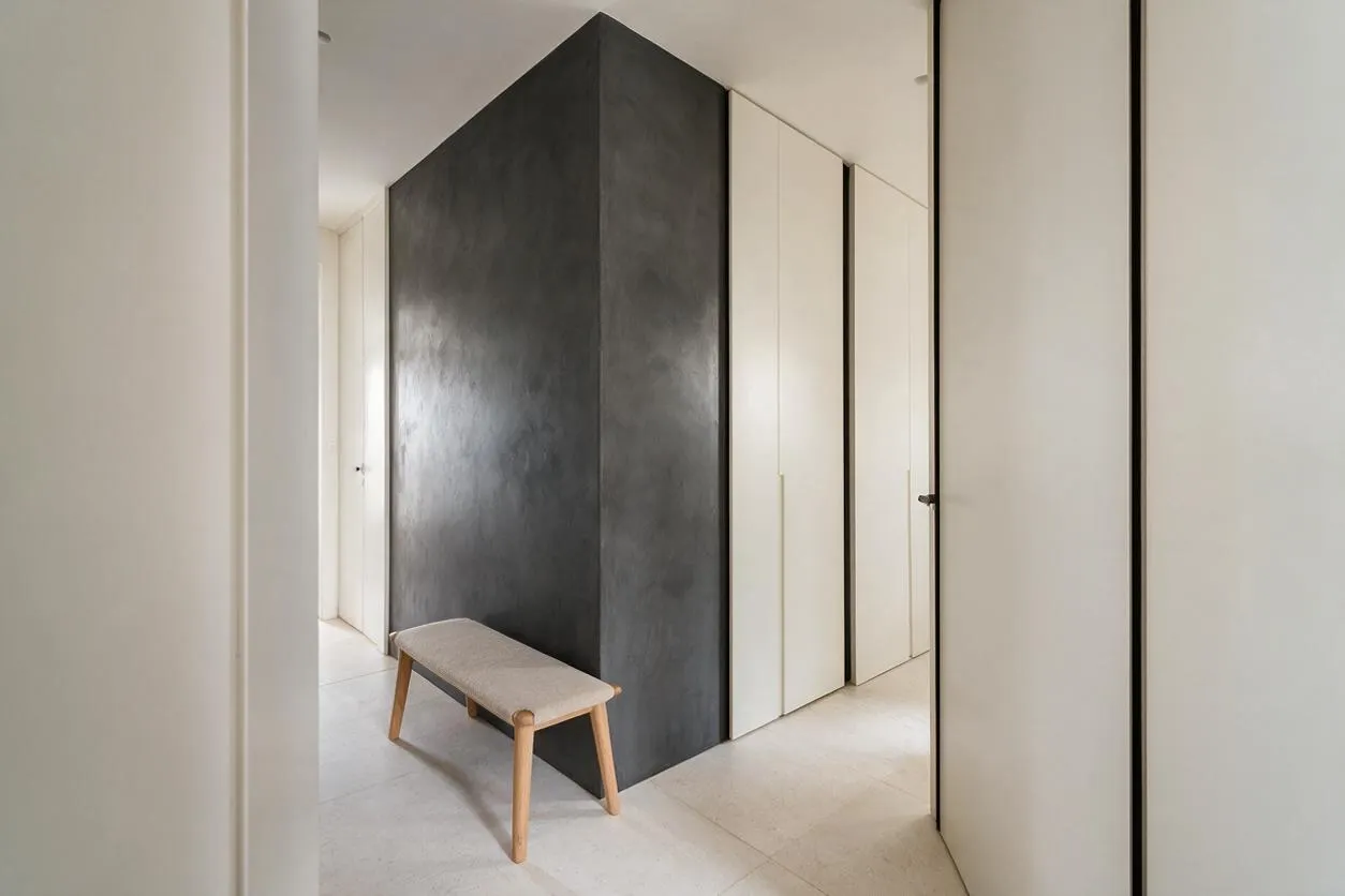 A minimalist entryway with smooth white walls, a tall charcoal-gray accent volume at the center, and light stone tile flooring. A small upholstered bench with pale wood legs sits in front, while flush white doors and slim black trim keep the space clean and calm.