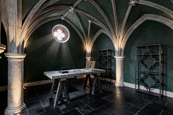 An atmospheric Gothic home office showcases a ribbed vaulted ceiling and limestone columns against deep green limewashed walls. Light streams through a round quatrefoil window onto a rough stone trestle desk, black slate flooring, and two blackened steel shelving units echoing the quatrefoil motif; subtle uplights wash the arches to create a cathedral-like mood around a minimalist workstation with a sleek computer and black leather swivel chair.