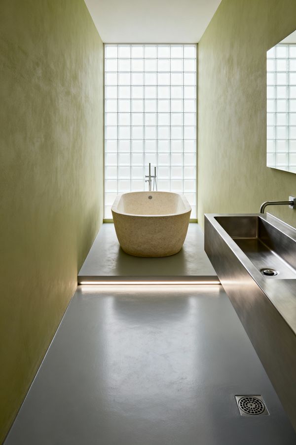 A narrow minimalist bathroom features muted olive plaster walls and a seamless gray floor leading to a raised platform with a freestanding oval limestone tub. A floor-mounted stainless tub filler stands before a wall of square glass blocks that diffuse daylight. At right, a long stainless-steel trough sink with a wall-mounted faucet aligns with a frameless mirror. A linear LED under the step and a discreet floor drain complete the clean, functional layout.