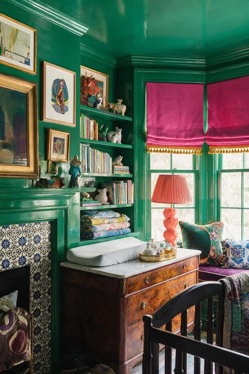 A maximalist nursery with glossy emerald-green walls, built-in shelves filled with children’s books, framed artwork, and small decorative animals. The room features magenta Roman shades, a coral lamp, a vintage wood changing dresser with a pale changing pad, patterned pillows, and a black crib in the foreground.