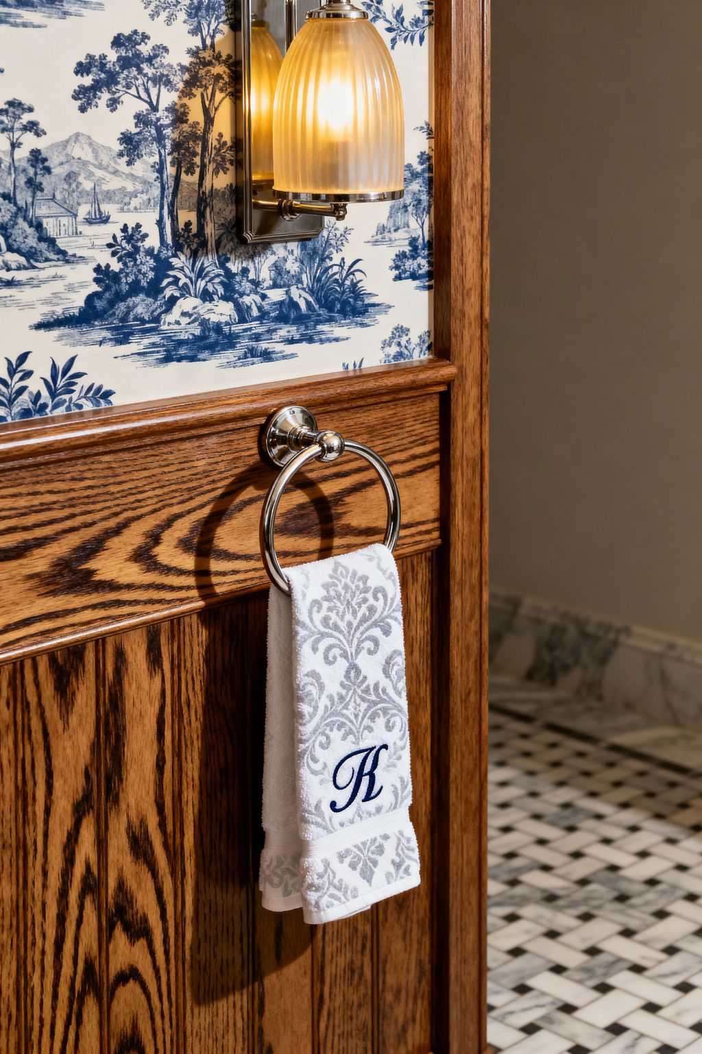 Close-up of a traditional bathroom shows stained oak wainscoting with a polished nickel towel ring holding a white hand towel patterned in soft gray damask and a navy monogram K. Above, a wall sconce with a ribbed glass shade casts warm light against blue-and-white toile wallpaper, while a black-and-white marble basketweave floor and marble base trim appear in the background.