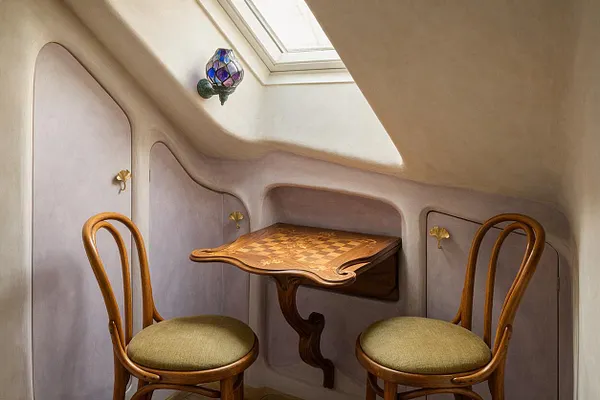 A small Art Nouveau game room nook under a sloped ceiling features a built-in wooden chess table with a carved pedestal base and two bentwood chairs with green upholstered seats. Soft mauve wall panels, brass floral hardware, and a stained-glass wall sconce create a warm, whimsical atmosphere.