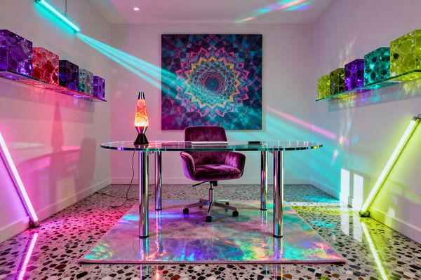 A psychedelic home office features a glass and chrome desk on an iridescent rug with a purple velvet swivel chair. A lava lamp sits on the desktop. Neon LED tubes in magenta, teal, and lime cast beams across white walls, terrazzo flooring, and a large kaleidoscopic mandala artwork. Floating glass shelves display translucent colored cubes, amplifying reflections for a vivid, prismatic workspace.