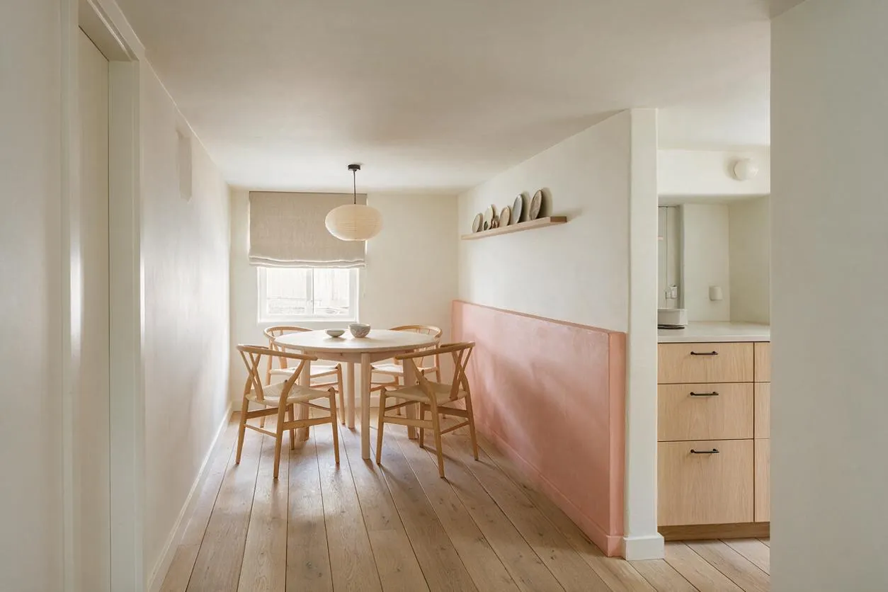 A narrow basement dining area with pale wood plank floors, white walls, and a round light wood table surrounded by matching Scandinavian chairs. A blush pink half-wall runs along one side, with a simple pendant light overhead, a linen window shade, and a small kitchen alcove visible to the right.
