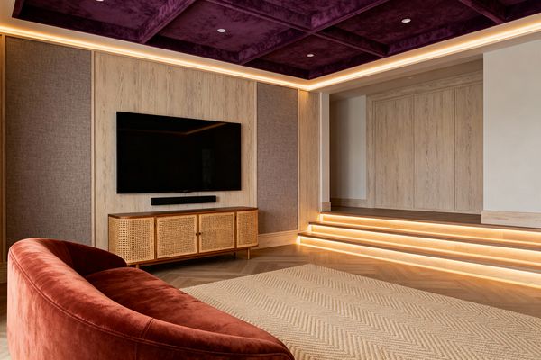 A transitional media room features a deep-plum upholstered coffered ceiling with warm LED cove lighting. A large wall-mounted TV sits on light oak panels flanked by gray fabric insets above a wood-and-cane media console and soundbar. A rust velvet curved sofa faces a pale herringbone rug over chevron wood floors. LED-lit steps lead to a raised platform with matching oak paneling, creating a plush, cinema-like atmosphere.
