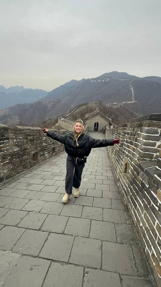 A person joyfully poses on the Great Wall of China, surrounded by scenic mountainous terrain.