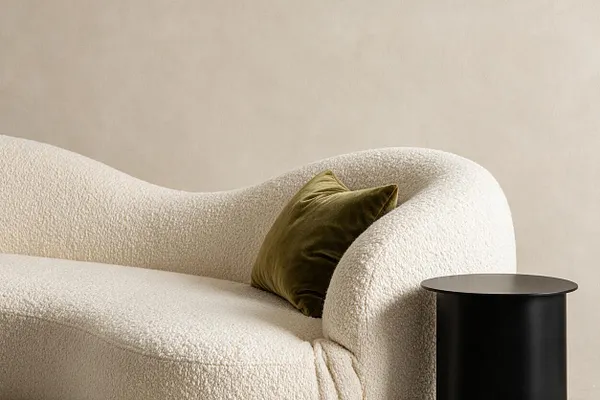 A modern seating vignette with a sculptural cream bouclé sofa featuring rounded arms and a matching seat, accented by an olive green velvet pillow. A small matte black cylindrical side table sits beside it against a warm beige wall.