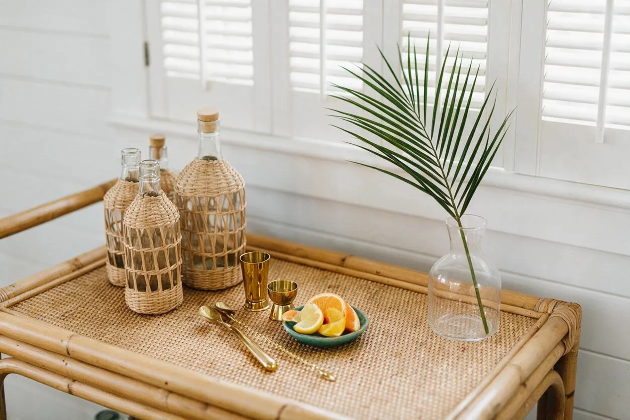 A tropical-inspired bar cart with a woven rattan top and bamboo frame holds glass bottles wrapped in natural fiber, gold cocktail tools, a small bowl of citrus slices, and a clear vase with a palm frond. White plantation shutters and soft daylight create a bright, airy backdrop.