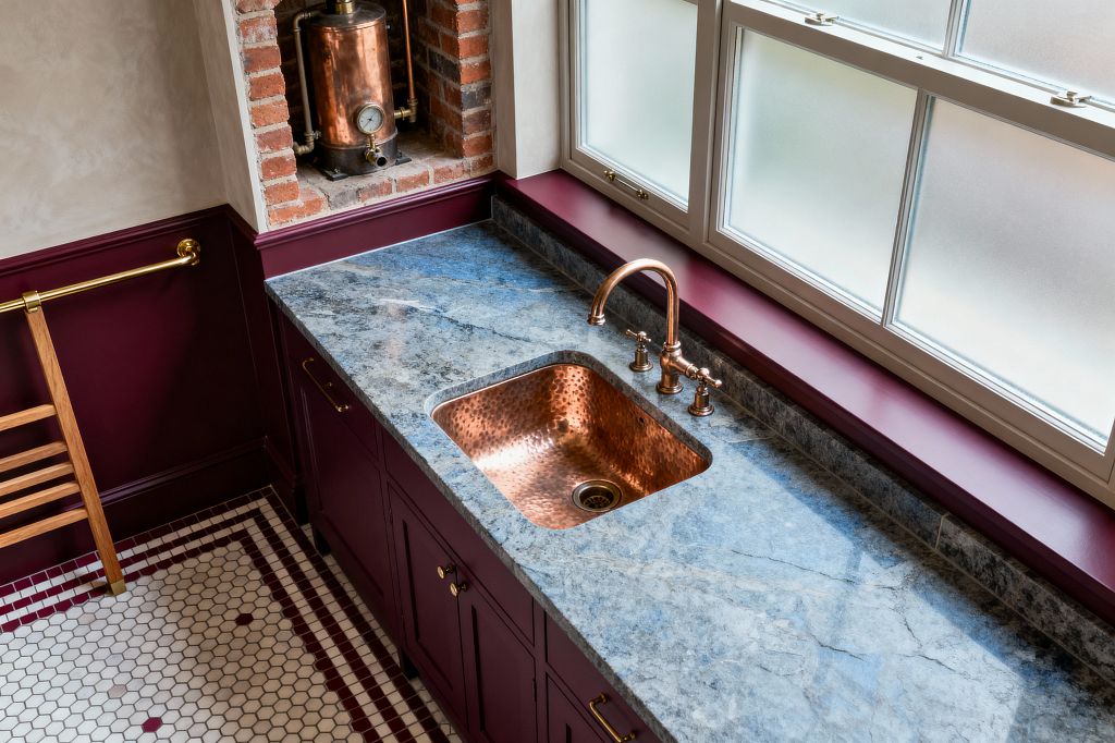 A Victorian-style laundry features burgundy paneled cabinetry topped with a blue-gray stone countertop, a hammered copper undermount sink, and a polished copper bridge faucet beneath large frosted sash windows. A recess of exposed red brick holds a vintage copper boiler, while white hex mosaic tiles with burgundy borders cover the floor, complemented by brass hardware and a wall rail for a richly detailed, period-appropriate workspace.