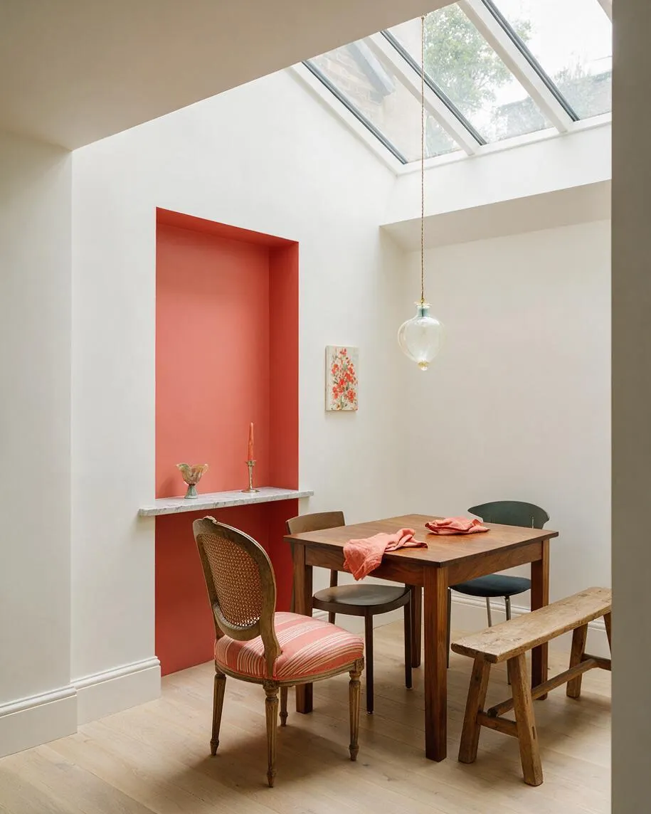A bright eclectic dining nook with a small wooden table and mixed seating under a skylight. White walls, a coral recessed alcove with a marble ledge, and a clear glass pendant create a warm, airy atmosphere.