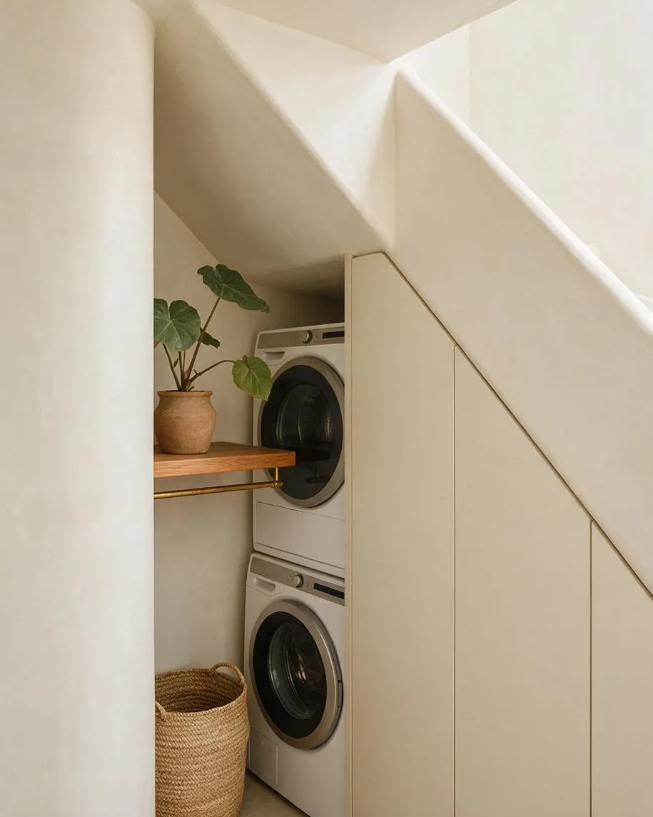 A small laundry room tucked beneath a staircase, with stacked front-loading washer and dryer behind smooth cream cabinetry. A warm wood shelf holds a terracotta pot with a leafy plant, and a woven basket sits on the floor, adding natural texture to the soft neutral space.