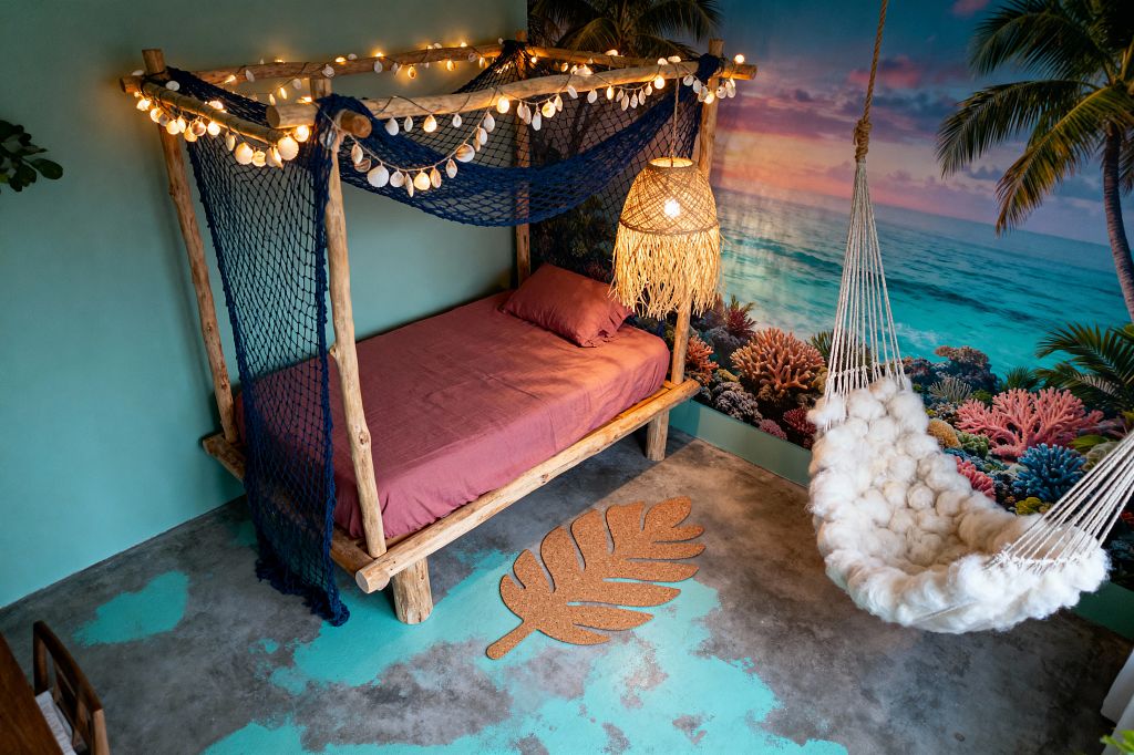 A kids’ room styled like a beach cabana features a rustic log-frame canopy bed draped with navy fishing net, shell garlands, and warm globe string lights. A woven rattan pendant lamp hangs over mauve-pink bedding. Opposite the bed is a white cotton-rope hammock with a fluffy wool seat. A large ocean mural with palm trees and coral reefs spans the wall, while a concrete floor with aqua paint accents and a cork leaf rug completes the tropical look.