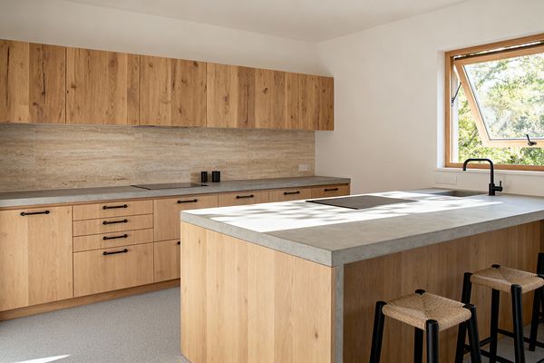 A bright Scandinavian kitchen with flat-front oak cabinets and a matching island topped in light gray concrete. A beige travertine backsplash runs the length of the back wall, matte black pulls and faucet add contrast, and three black stools with woven rush seats line the island beneath a wide wood-framed window bringing in natural light.