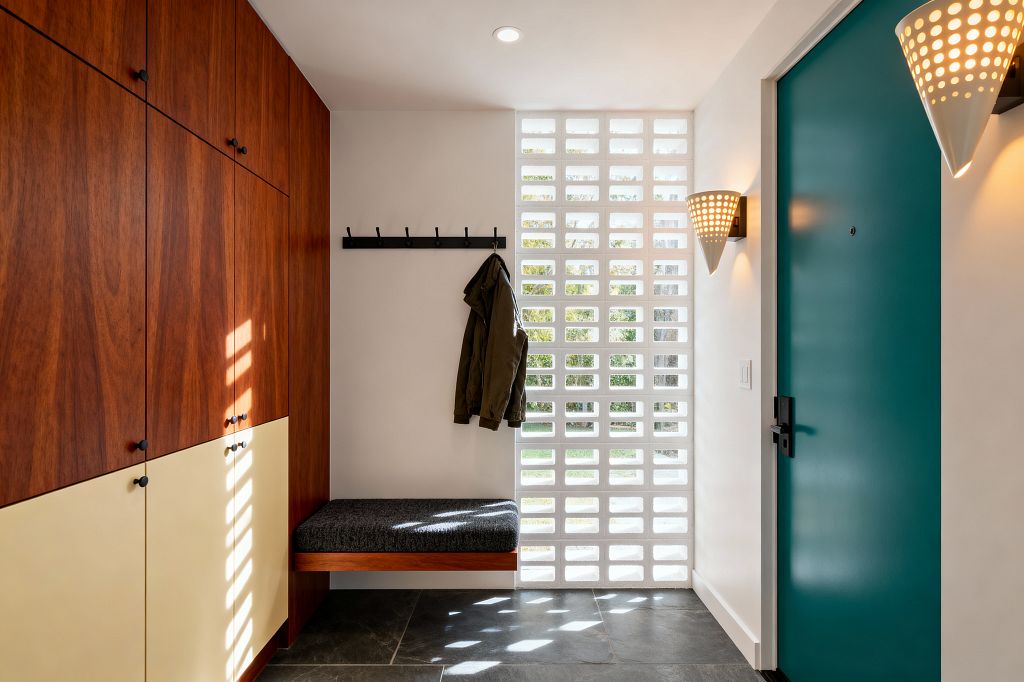 A mid-century modern mudroom with tall walnut-toned cabinets and cream laminate base doors at left, a floating bench with a charcoal tweed cushion, and black metal wall hooks. At right, a teal entry door sits beside a white breeze-block screen that filters sunlight onto dark slate tile. Perforated cone sconces cast warm light against crisp white walls.