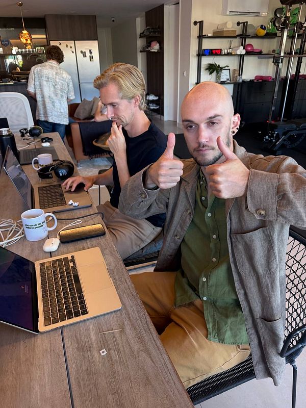 Two men working at a shared wooden table in a modern coworking space, with one man giving a double thumbs-up.