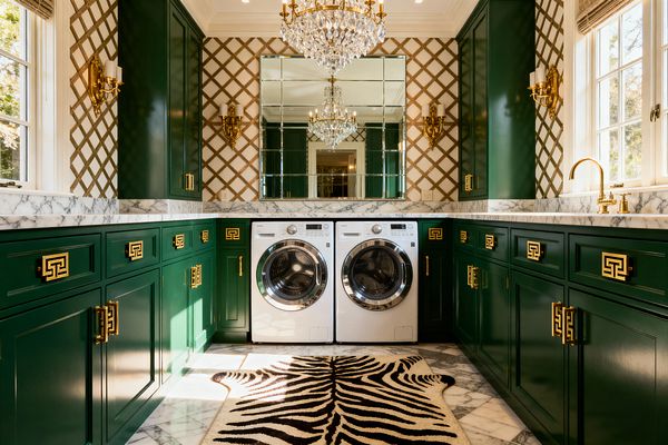 A glamorous laundry room with emerald green cabinets and brass Greek key pulls forms a U-shaped workspace around a front-loading washer and dryer, topped with white marble counters and a gray-veined marble floor. A mirrored backsplash reflects a crystal chandelier and brass double sconces, while cream-and-gold trellis wallpaper, a zebra-print rug, a window with a woven Roman shade, and a brass gooseneck faucet complete the scene.