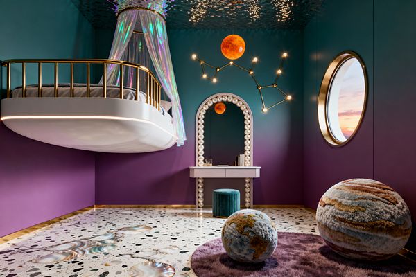 A kids’ bedroom with teal-to-plum ombré walls features a floating white bed with a brass rail and an iridescent canopy on the left. Centered is an arched vanity edged with large pearl-like bulbs and a teal velvet stool, with a brass constellation light and an orange planet pendant below a rippled reflective ceiling; a brass-trimmed porthole window sits on the right, and a terrazzo floor with shell-like inlays holds a lilac shag rug and plush planet poufs.