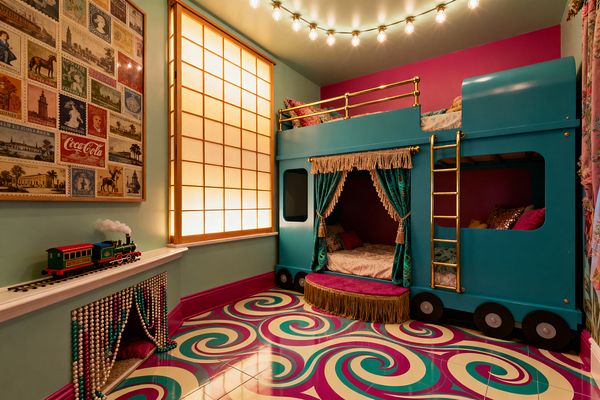 A children's room in maximalist style features a teal train-shaped bunk bed with brass ladder and rails, velvet teal curtains with gold fringe, and a magenta step. A glossy floor of cream, teal, and fuchsia swirls anchors mint walls with a framed vintage stamp collage, a toy train on a shelf above a beaded-curtain nook, and warm light from a backlit shoji panel and ceiling globe string lights.