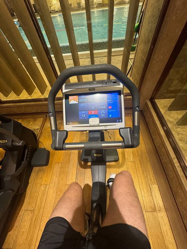 A person is using a stationary bike in a gym with a view of a swimming pool.