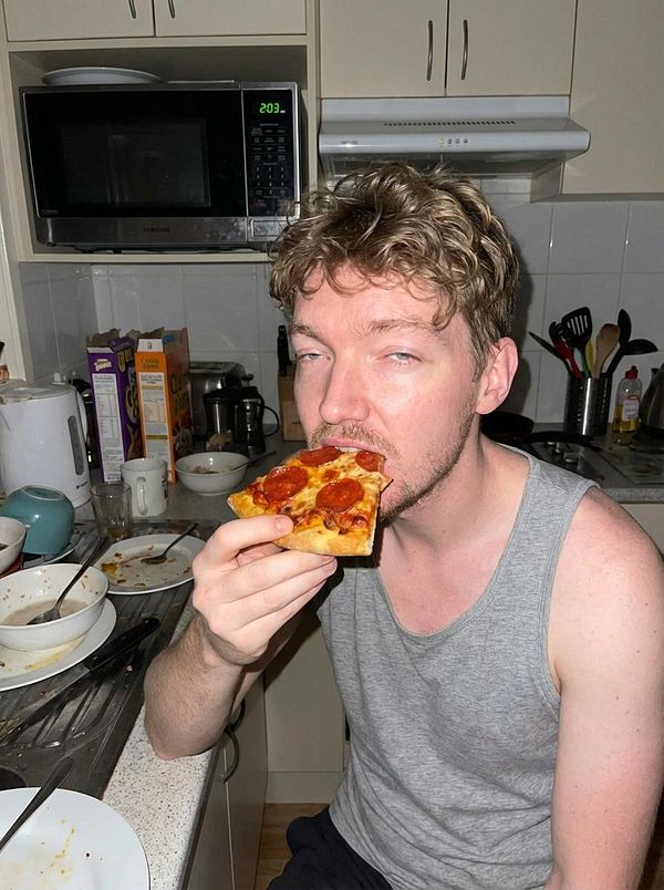 A person is sitting in a messy kitchen, holding a slice of pepperoni pizza.