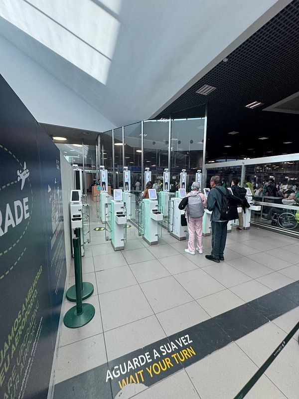 The image depicts a queue at the new electronic passport gates at Lisbon Airport, highlighting a frustrating experience for travelers.