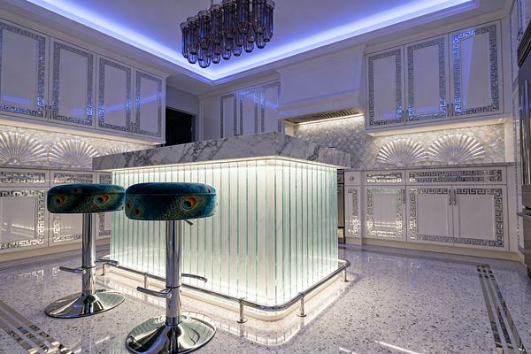 A glamorous kitchen centers on a glowing fluted glass island capped with white marble and edged in chrome, accompanied by two teal bar stools upholstered in peacock-pattern velvet on chrome pedestals. High-gloss white cabinets with mirrored Greek key borders and an iridescent scallop backsplash with shell-fan reliefs shimmer under blue cove lighting, with a smoked glass chandelier above and a polished terrazzo floor below.