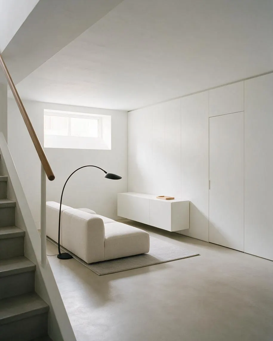 A bright minimalist basement lounge with white walls and a low white sofa on a small rug. A black arched floor lamp stands beside the seating, while a long wall of seamless built-in cabinets and a small horizontal window keep the space clean and uncluttered. 