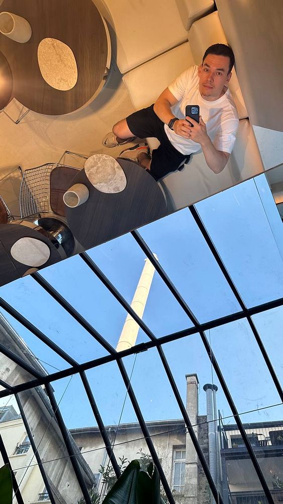 A person takes a selfie while sitting in a modern, stylish café with a glass ceiling showing a blue sky.