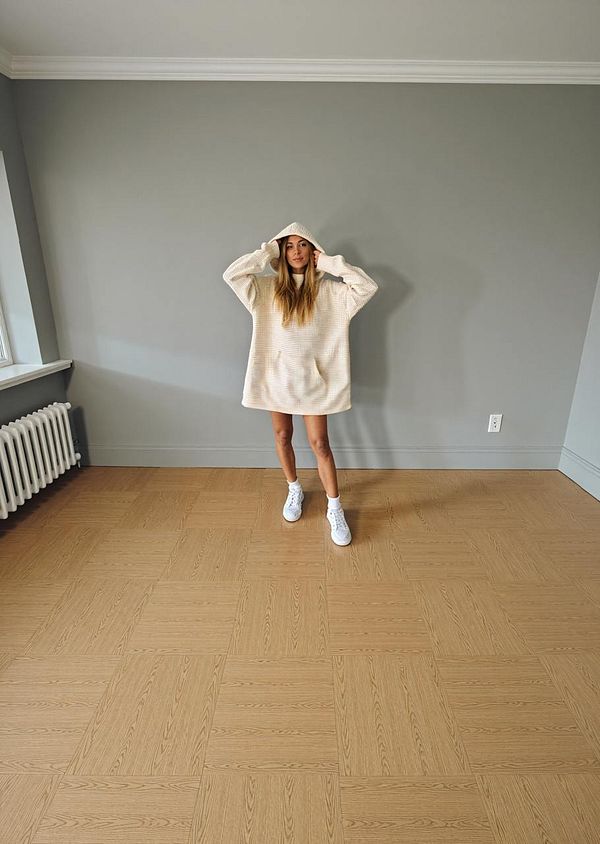 A young woman stands in a spacious, empty room wearing a large, light-colored hoodie and white sneakers.
