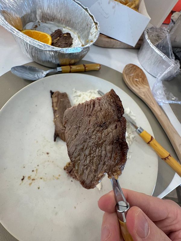 A hand holds a piece of well-cooked picanha meat above a plate with rice.