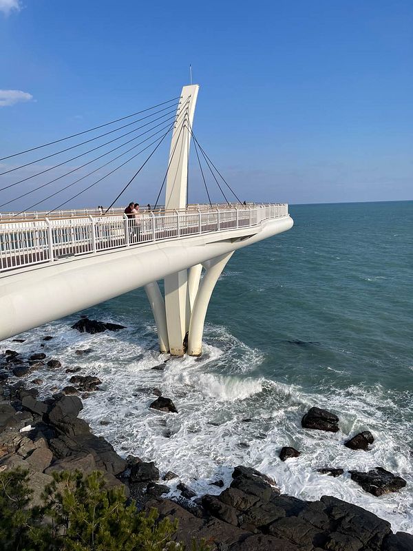 A modern bridge extends over rocky shores with waves crashing below in Busan.