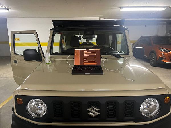 A custom-modified Suzuki Jimny SUV is parked on a dirt path in a lush, green forested area.