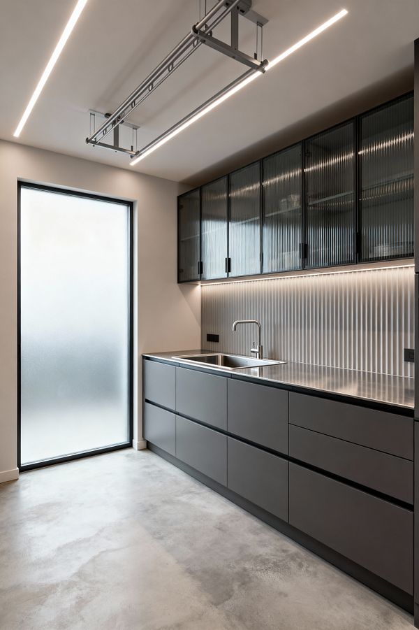A modern laundry room with matte charcoal base drawers, a stainless-steel countertop, and a square sink beneath black-framed upper cabinets with reeded glass. A vertical fluted light-gray backsplash glows with continuous under-cabinet LEDs. Linear recessed ceiling lights and a ceiling-mounted drying rack span the space. At the left, a full-height frosted glass door diffuses daylight onto a smooth gray polished concrete floor, creating a clean, minimalist atmosphere.