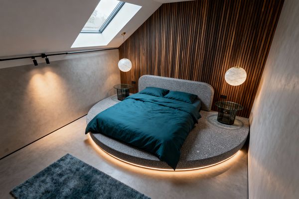 A contemporary attic bedroom features a round, speckled gray terrazzo platform bed with a curved headboard and deep teal bedding. A concealed LED strip casts a warm halo beneath the platform. A vertical walnut slat wall spans behind the bed. Two marble-like alabaster globe pendants hang above circular glass nightstands with black bases. Smooth gray microcement walls, black track lighting, a skylight for daylight, and a blue-gray shag rug complete the minimalist space.