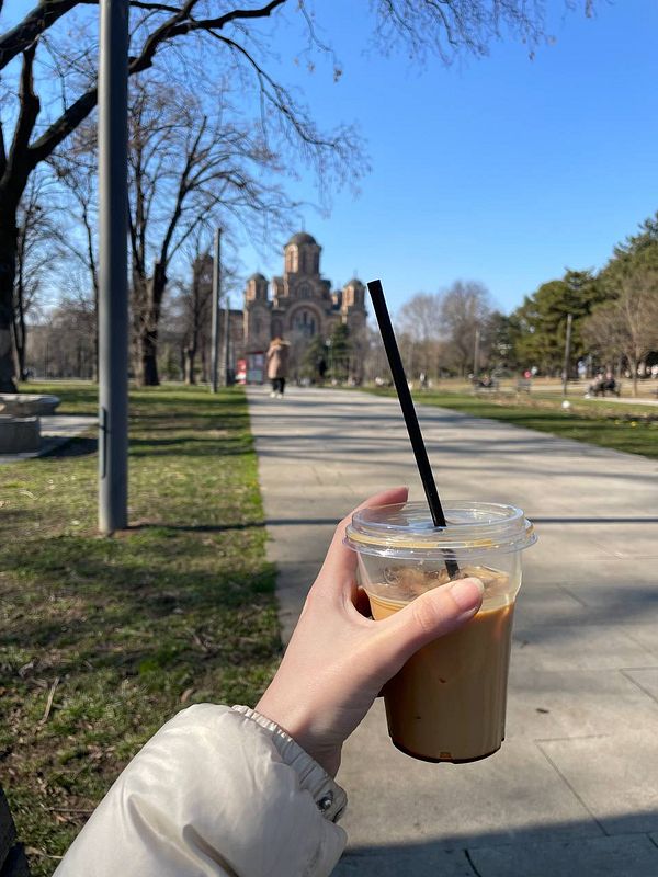 A hand holding an iced coffee in a park setting with a church in the background.