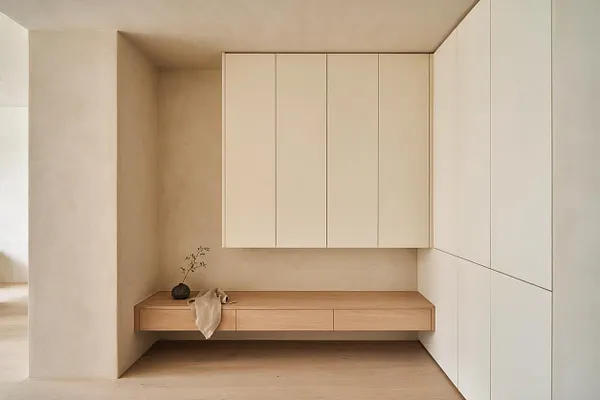 A minimalist entryway with a floating light wood bench built into a recessed alcove, topped by a small vase with a branch and a draped neutral cloth. Large matte cream cabinetry fills the wall above and to the right, while soft beige walls and pale wood flooring create a calm, seamless look.