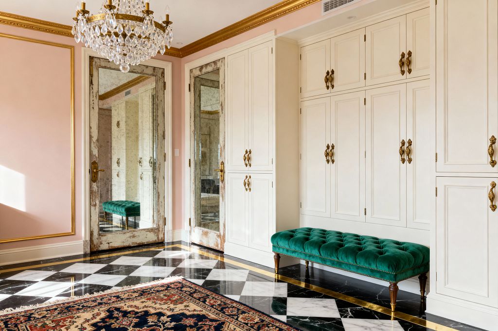 A mudroom with blush pink walls framed by gilded molding features floor-to-ceiling cream cabinetry with paneled doors and antique brass pulls. Two antiqued mirrored doors and a crystal chandelier reflect natural light onto a polished black-and-white marble checkerboard floor edged with brass; in front sits a tufted emerald velvet bench with turned wood legs and nailhead trim, while a Persian-style rug adds pattern near the entry.