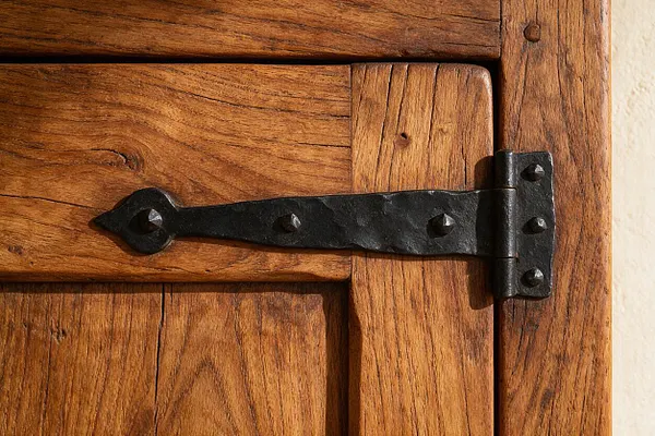 Close-up of a rustic wooden door or cabinet with varied plank construction and a black hand-forged iron strap hinge fastened with visible rivets. The warm wood grain and dark metal create a traditional Southwestern look, with a plaster wall visible at the edge.