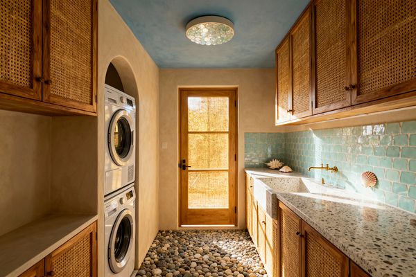 A narrow coastal laundry room with sand-colored plaster walls and a sky-blue ceiling lit by a round, shell-like flush mount. Warm wood cabinets with woven rattan doors line the right wall, opposite an arched niche housing a stacked white washer and dryer. A speckled terrazzo counter with a deep integrated sink sits beneath glossy seafoam tiles and a brass wall-mounted faucet. River-pebble flooring leads to a wood-framed glass door screened with woven reed and glowing with sunlight.