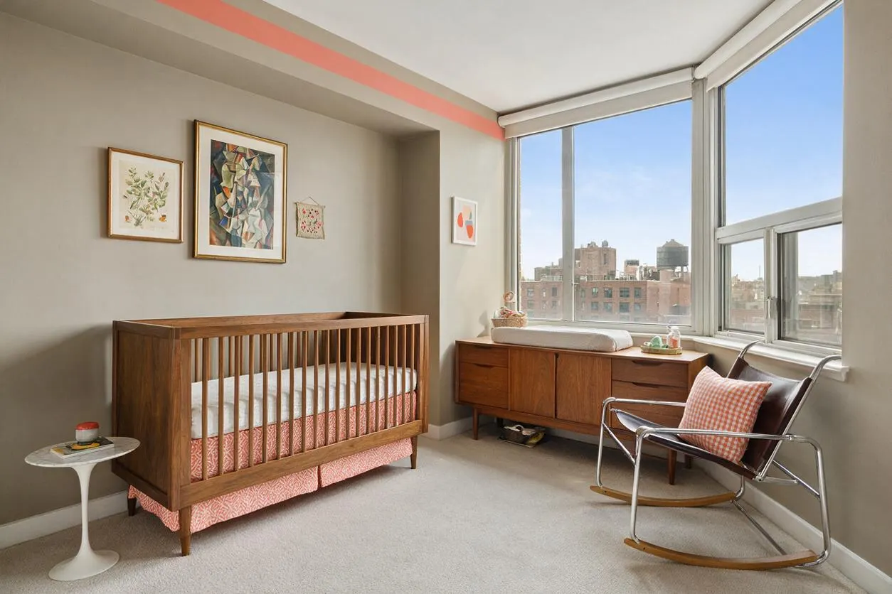 A bright nursery with a warm wood crib against a soft beige wall, framed artwork above it, and a long wooden dresser beneath large corner windows. A chrome and leather rocking chair sits by the window, while a small side table and pink patterned crib skirt add playful contrast.