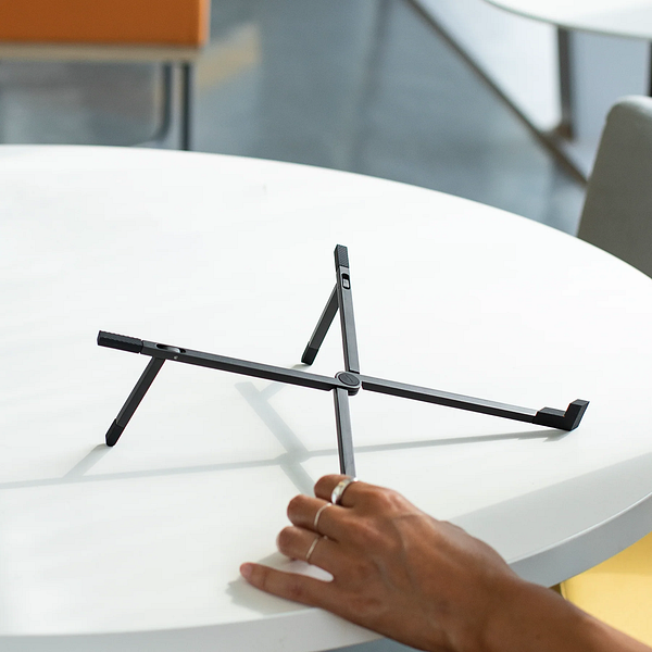 A lightweight laptop stand is positioned on a table with a hand reaching towards it.