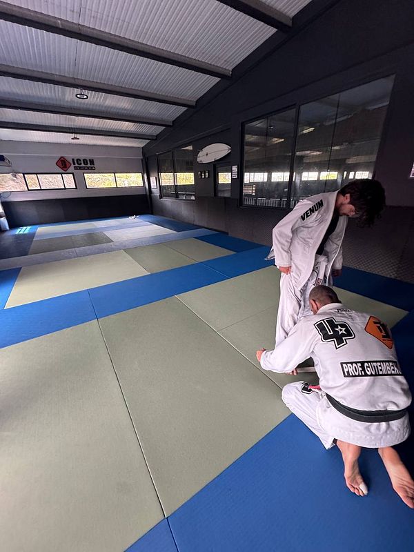 Two practitioners are preparing for Brazilian Jiu-Jitsu training on a mat in a gym.